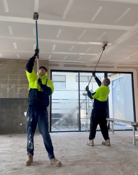 Two plasterers coating the ceiling joints in a new build, working overhead with taping tools on extension handles.