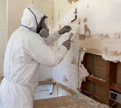 Wall lining being stripped out in full protective equipment in an older bathroom, before new sheeting goes back on.