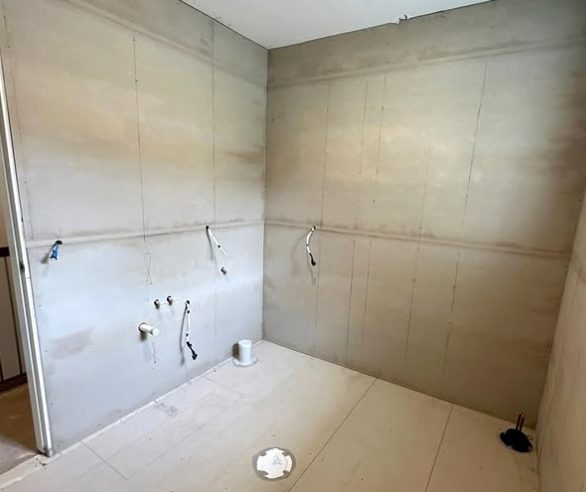 A bathroom sheeted in water resistant board, set and sanded, with the tap and shower rough ins left exposed for the plumber.