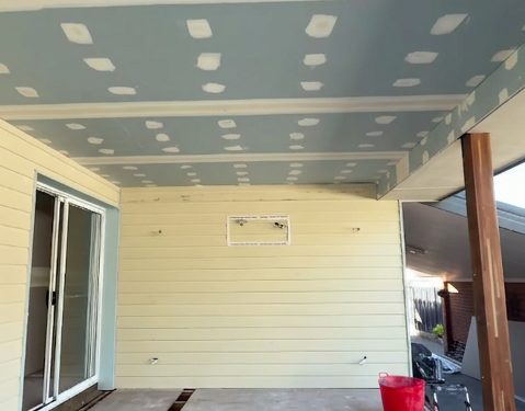 A patio ceiling lined and set under the main roofline, above a weatherboard wall and a sliding door.