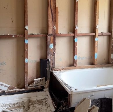 Wall lining stripped back to the studs beside a bath after storm damage, with the wet board removed.